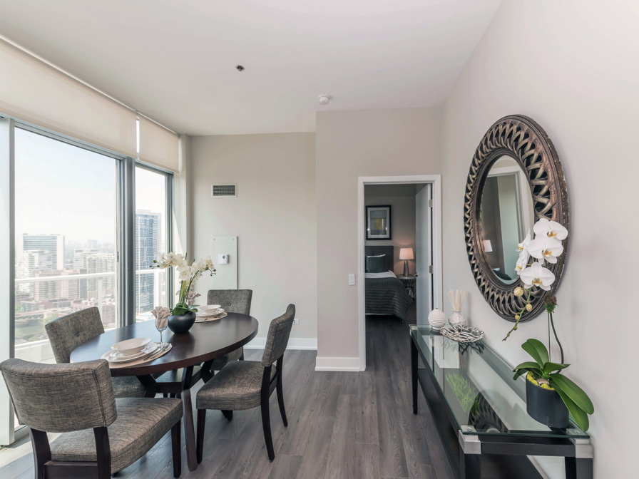 Modern dining area with a round table set for four, large windows revealing a city view, and a glass console table with decorative items and a mirror on the wall. Bedroom visible through the doorway.