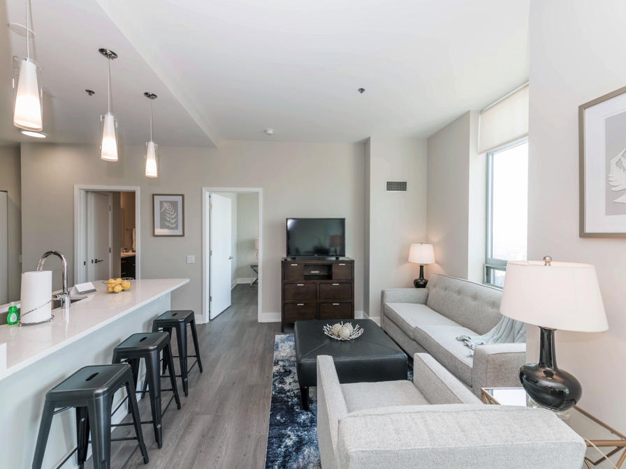 Modern apartment living area with a kitchen island, bar stools, couches, a rug, a cabinet with a TV, and a table lamp. Walls are light-colored, and the flooring is dark wood.