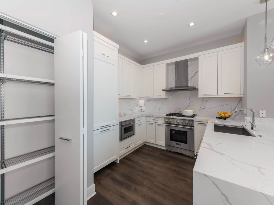 Modern kitchen with white cabinets, marble countertops, stainless steel appliances, and an open pantry. Dark wood floor contrasts with light decor. Hanging light fixtures provide illumination.