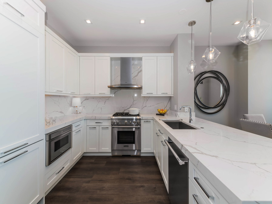 Modern kitchen with white cabinetry, marble countertops, stainless steel appliances, and hanging pendant lights. Dark wood flooring contrasts with light gray walls and circular wall decor.