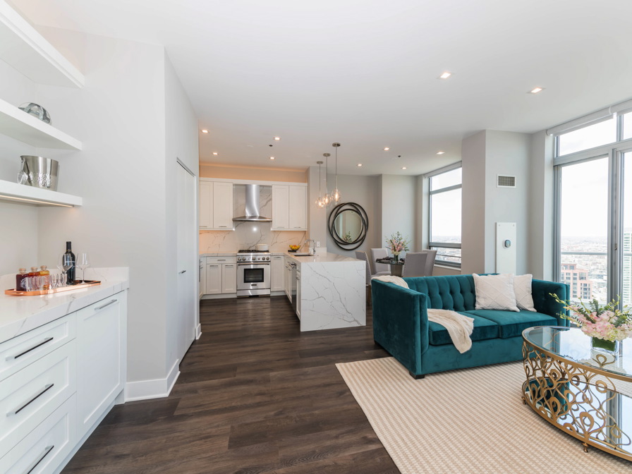 Modern open-concept kitchen and living area with white cabinets, stainless steel appliances, teal sofa, glass coffee table, and large windows with city views.
