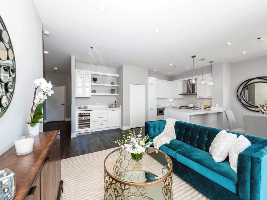 Modern living room and kitchen area with a teal sofa, glass coffee table, and white cabinetry. Decorative mirrors and pendant lights enhance the bright, open space.