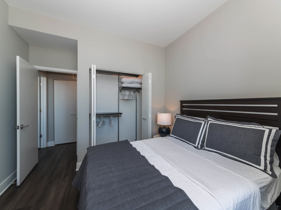 Modern bedroom with a neatly made bed, gray and white bedding, an open closet with shelves, and a nightstand with a lamp.