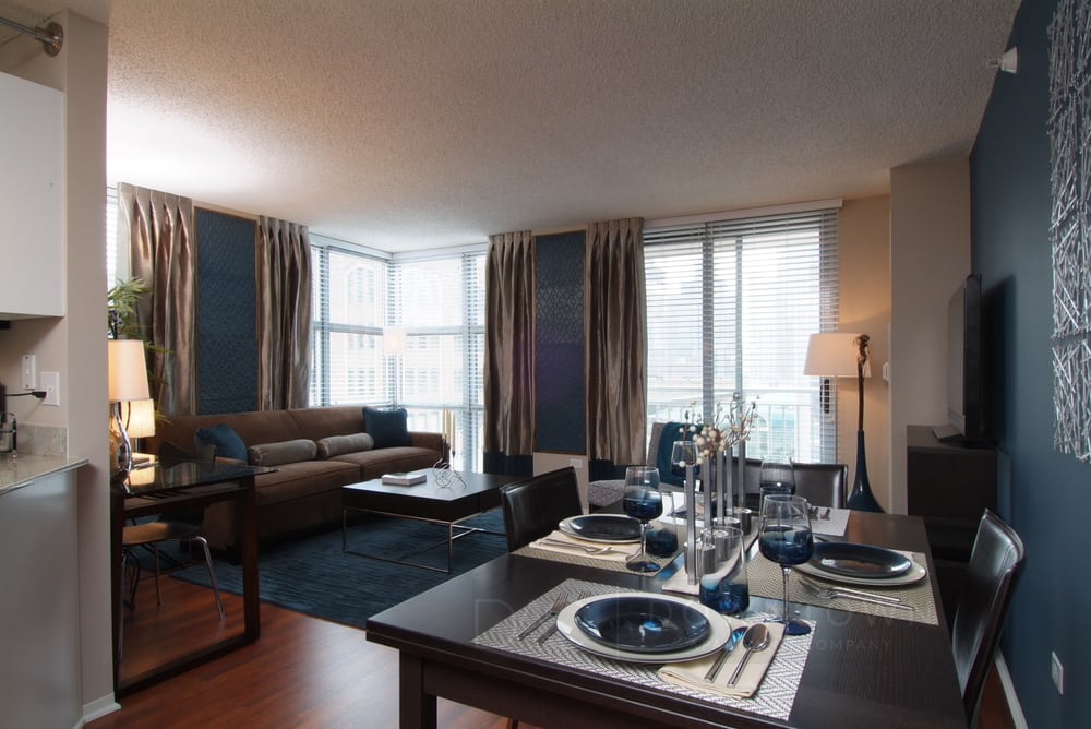 Modern dining and living area with a dark wood table set for four, navy accents, a corner sofa, and large windows with beige curtains.