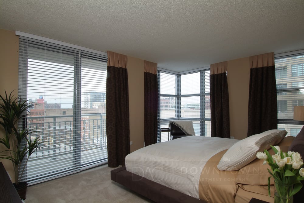 Bedroom with large windows, beige walls, and brown curtains. Features a bed with white linens, plants, and a cityscape view outside.
