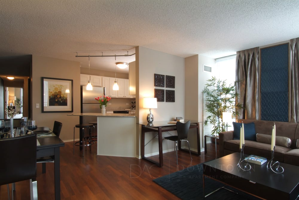 Modern living room with hardwood floors, a workspace, and a view into a kitchen with hanging lights. A large window with curtains provides natural light.