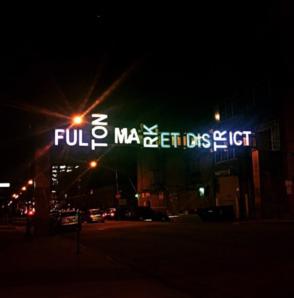 Neon sign reading "Fulton Market District" illuminated at night above a city street.