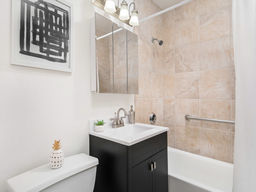 Modern bathroom with beige-tiled shower and white curtain. Dark vanity with white sink, and a framed abstract print. Wall-mounted mirror cabinet above sink. White toilet with decorative pot.