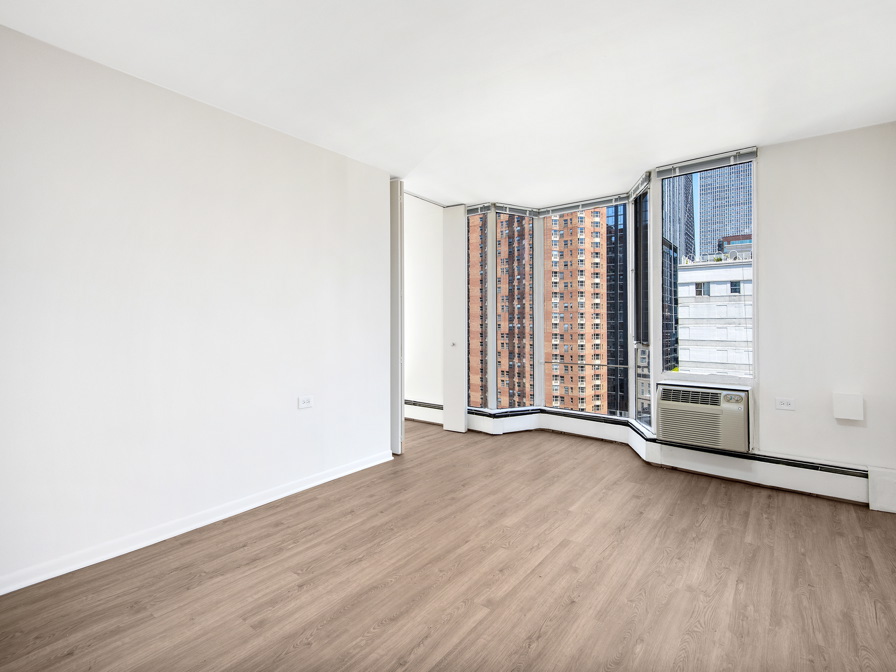 Empty room with wood flooring, large windows, and a city view. Walls are white, and there's a window air conditioning unit installed.