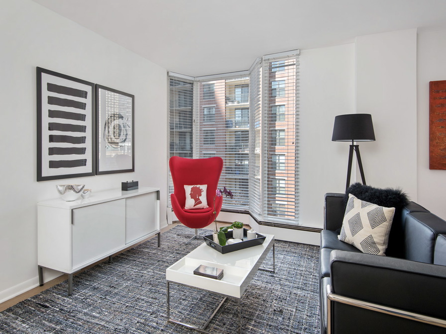 Modern living room with a red chair, black sofa, white coffee table, and floor lamp. Large windows with blinds, abstract artwork, and minimal decor.