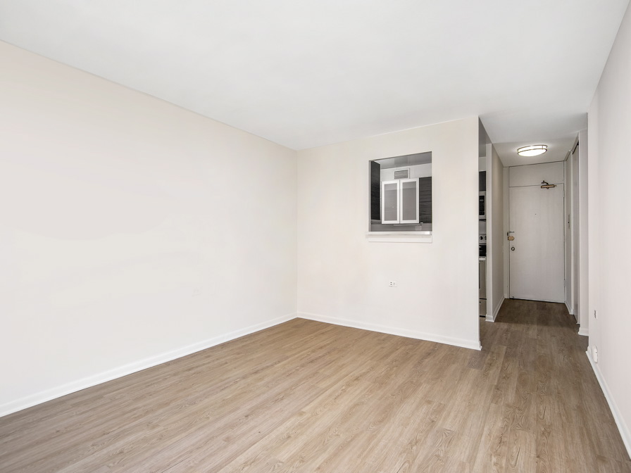 Empty room with light wood flooring, white walls, and a rectangular window. A narrow hallway on the right leads to a door.
