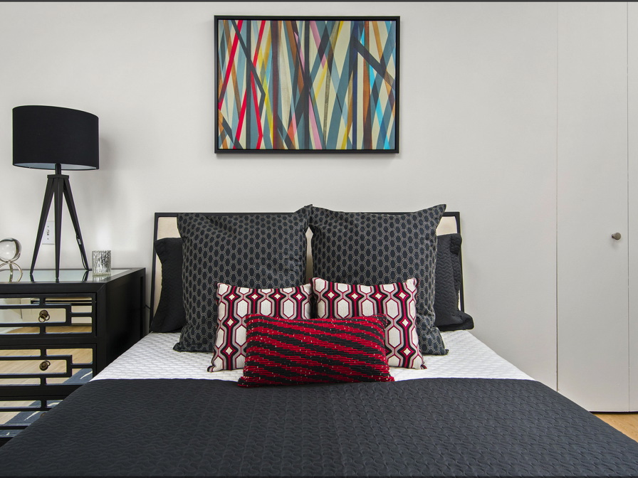 Modern bedroom with a black and white bed, geometric-patterned pillows, and a colorful abstract painting above. A black lamp sits on a bedside table with drawers.