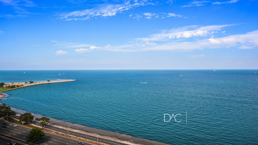 Aerial view of a large body of water with a curved shoreline and road on the left. The sky is clear with scattered clouds.
