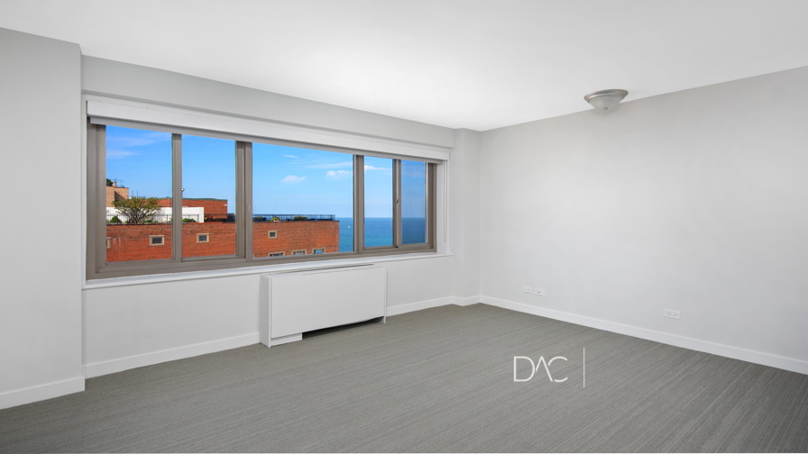 Bright, empty room with large window showing a view of red brick buildings and the ocean under a blue sky. Neutral walls, gray carpet, and a ceiling light fixture are visible.