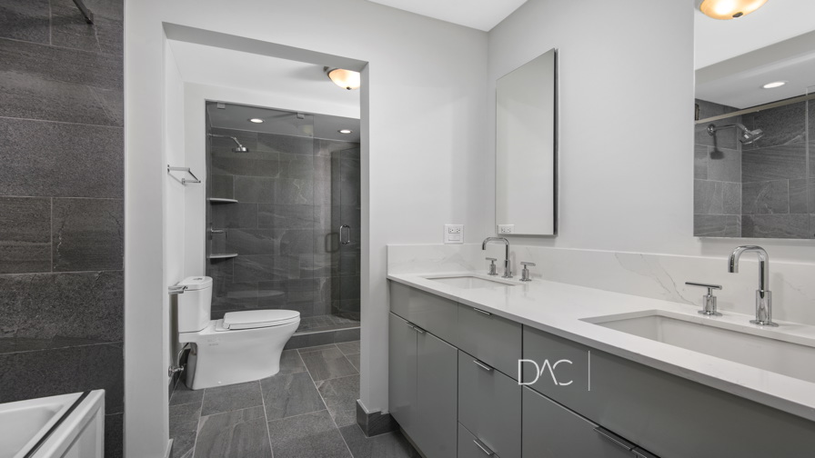Modern bathroom with gray tiles, double sinks, mirrors, and a glass-enclosed shower. A toilet is next to the shower. White walls and ceiling lights enhance brightness.
