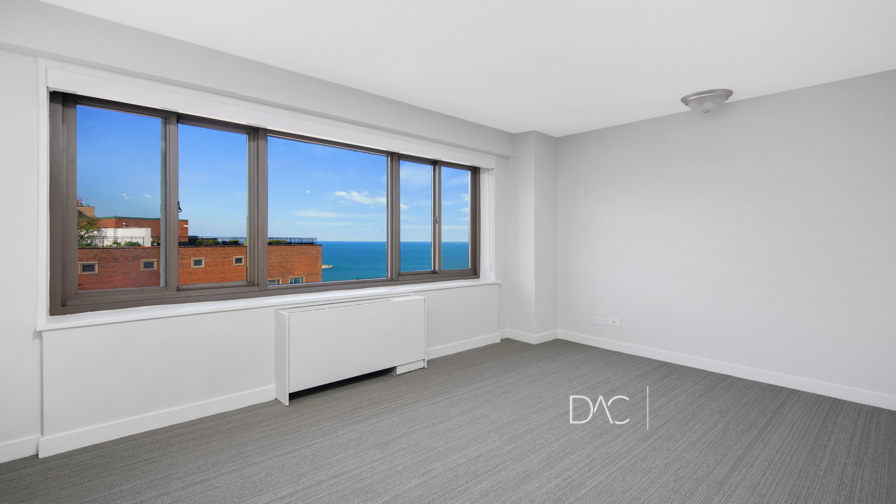 Empty room with large window overlooking the ocean and nearby buildings. Room has gray carpet, white walls, and ceiling light fixture.