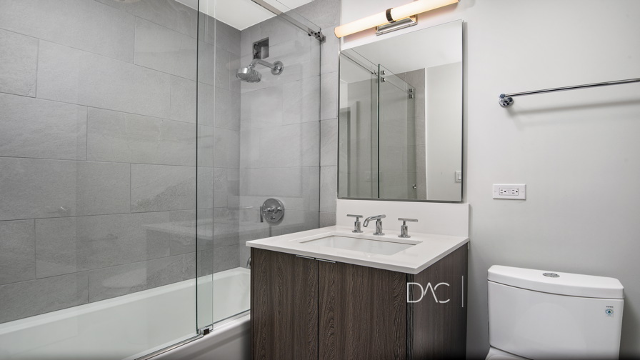 Modern bathroom with a bathtub-shower combo, glass door, wall-mounted mirror, sink with a wooden cabinet, and a toilet.