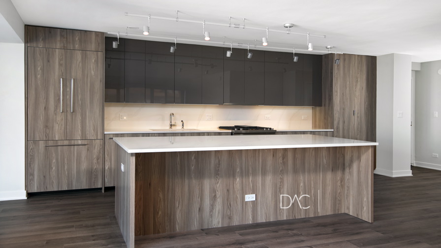 Modern kitchen with dark wood cabinets, glossy upper cabinets, a built-in oven, and a large island with a white countertop under track lighting.
