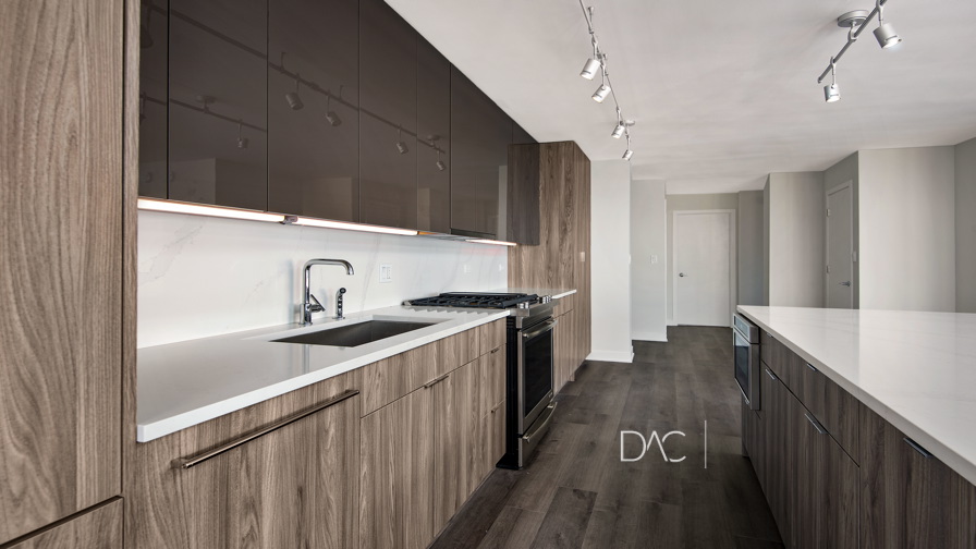 Modern kitchen with wooden cabinets, white countertops, stainless steel sink, and built-in oven. Overhead track lighting illuminates the space.