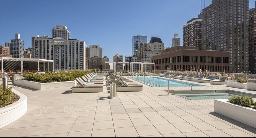 Rooftop pool area with lounge chairs and planters, surrounded by tall city buildings under a clear blue sky.