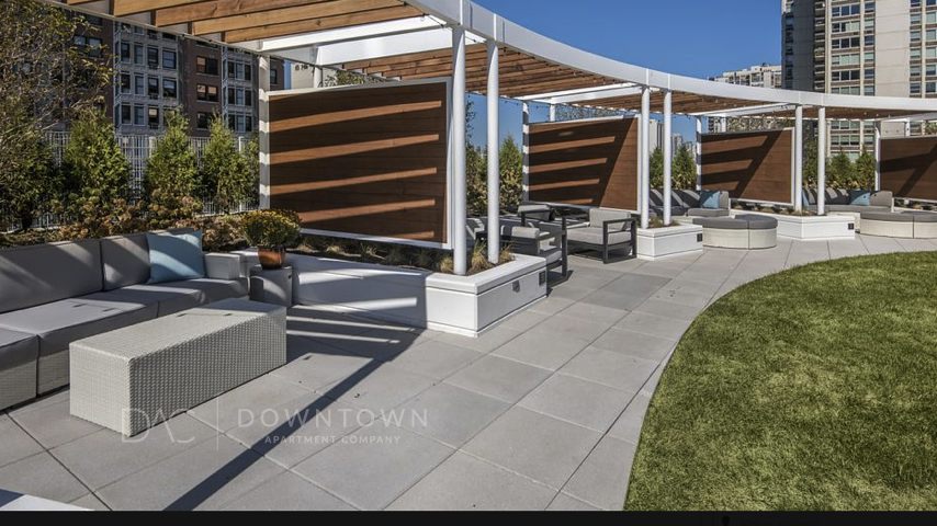 Outdoor rooftop lounge area with modern seating, wooden dividers, and a grassy section, surrounded by city buildings under a clear blue sky.