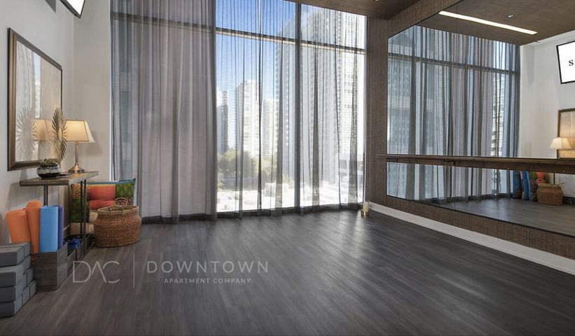 A bright room with large windows, dark wood floors, exercise equipment, and a mirror. City buildings are visible outside.