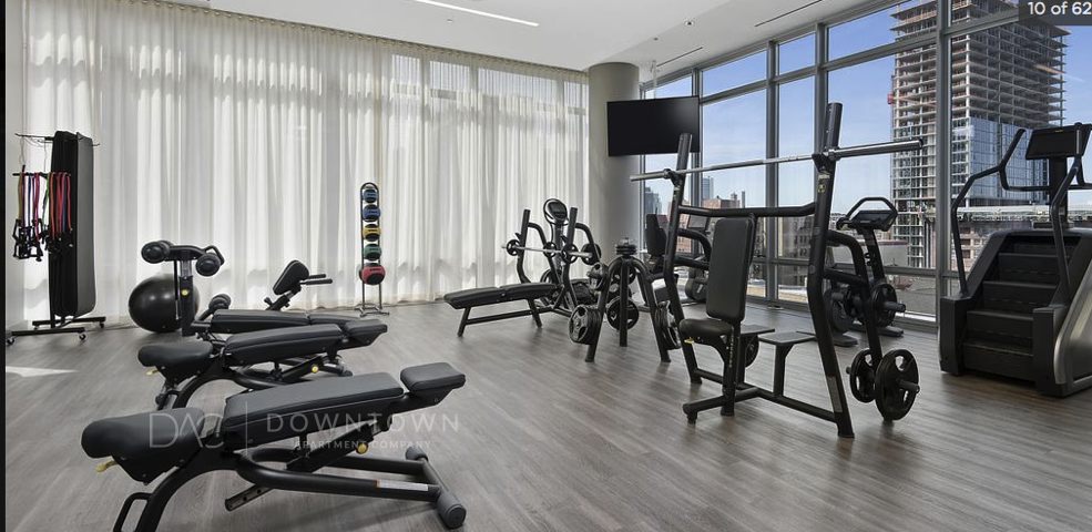 A modern gym with exercise equipment, including weight benches, stationary bikes, and a treadmill. Large windows provide natural light and a city view.