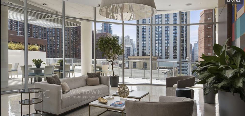 Spacious modern living room with large glass windows showcasing a city skyline. Features neutral-toned furniture, a potted plant, and access to a balcony with outdoor seating.