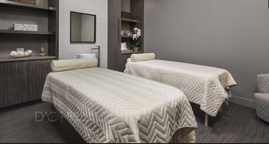 Two massage tables with cream-colored blankets in a spa room, featuring wooden shelves, a mirror, and a sink.