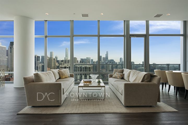 Spacious living room with two beige sofas, a coffee table, floor-to-ceiling windows, and a city skyline view. Light wood flooring and modern decor.