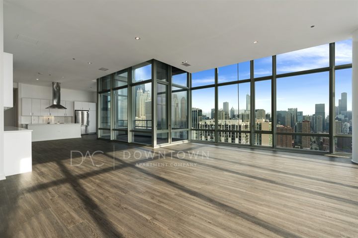 Spacious modern apartment with large windows offering a city skyline view. Features an open-plan layout with a kitchen and wooden flooring.