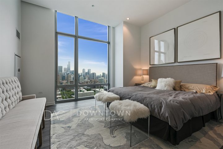 Bedroom with a large bed, two abstract paintings, and a bench. Floor-to-ceiling window offers a city skyline view. Neutral tones dominate the decor.