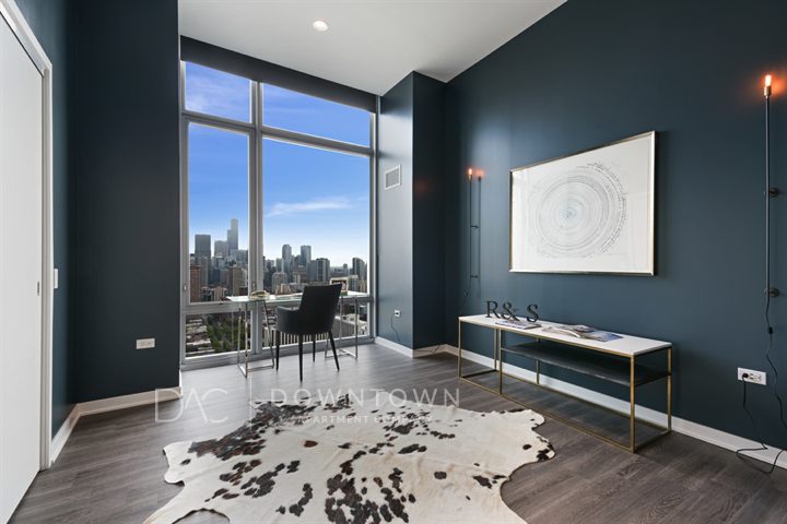 A modern room with dark teal walls, tall windows showcasing a cityscape, a cowhide rug, and a minimalist desk with a book and art piece on the wall.