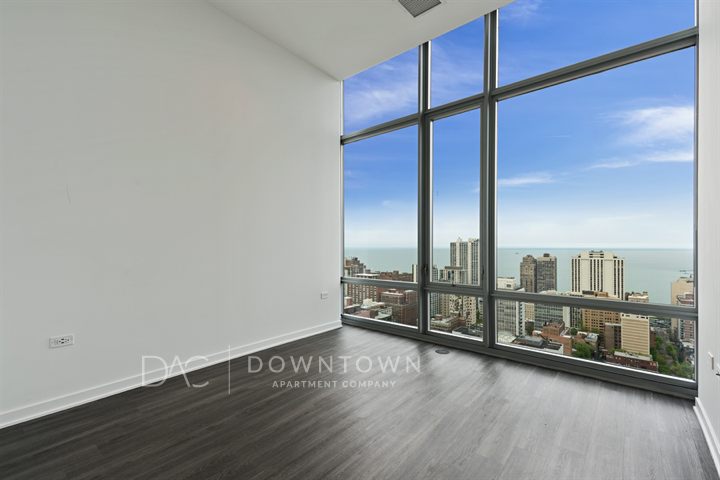 High-rise apartment with floor-to-ceiling windows showcasing urban skyline and lake view. Empty room with dark wood flooring and white walls.