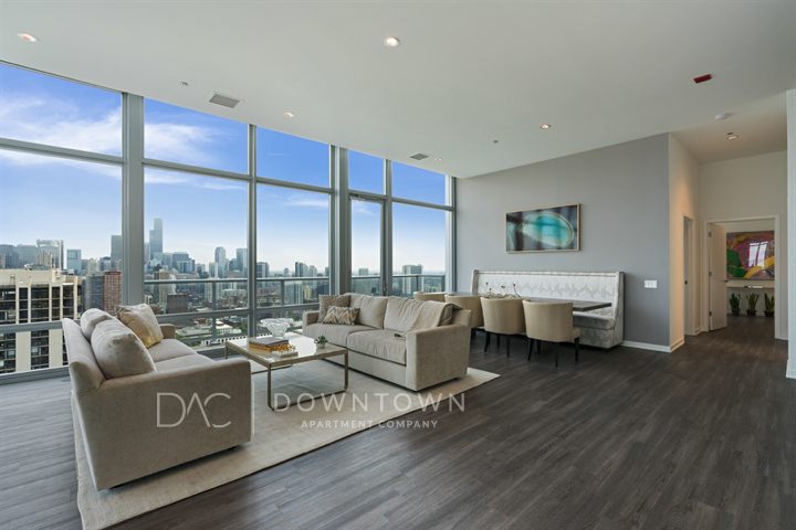 Spacious modern living room with large windows, city skyline view, light gray sofas, a glass coffee table, and a dining area with a bench and chairs.