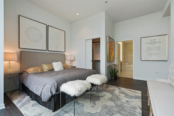 Modern bedroom with a large bed, gray bedding, and two cushioned stools. Wall art, lamps, and a rug complement the decor. A doorway leads to a bathroom with marble tiles.