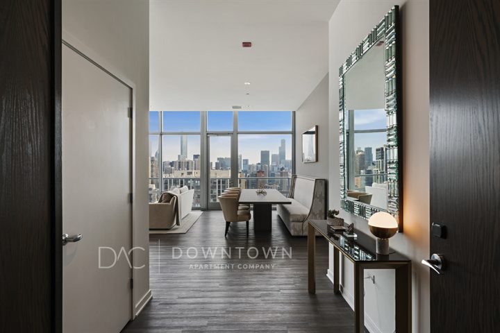 Modern apartment entrance leading to a living area with large windows showing a city skyline. Decor includes a mirror, console table, and seating, all in neutral tones.