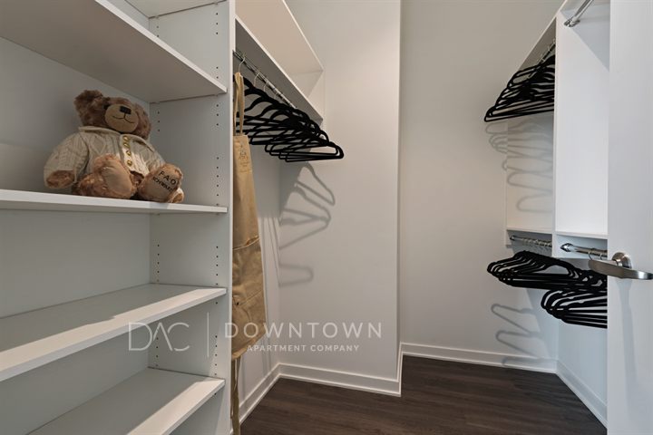 A spacious walk-in closet with empty shelves, hanging racks with black hangers, and a teddy bear. Floor is dark wood.