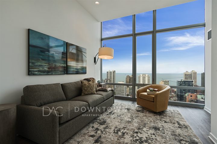 Modern living room with floor-to-ceiling windows, gray sofa, brown armchair, abstract wall art, and a view of city skyline and sea.