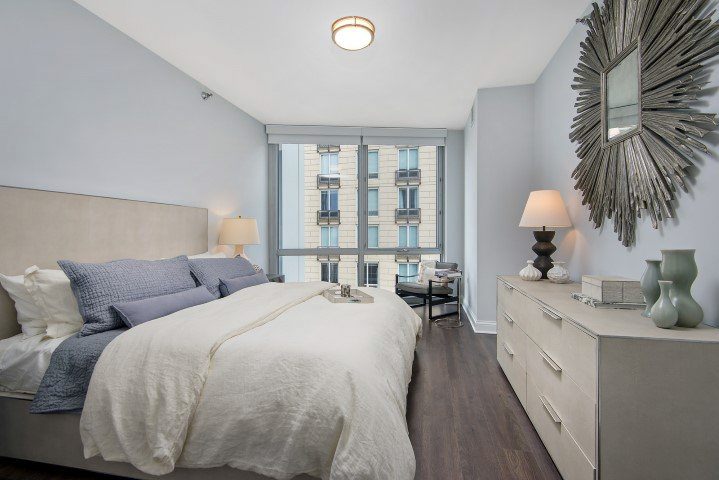 Modern bedroom with a large bed, grey and white bedding, two bedside lamps, a sunburst mirror, and a dresser. Floor-to-ceiling window reveals a building exterior.