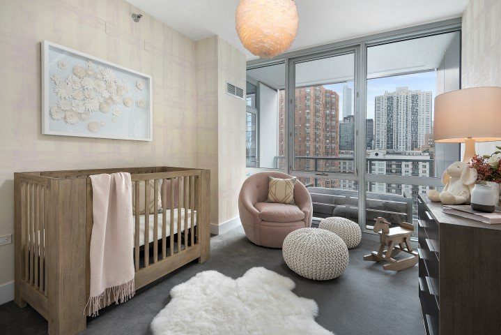 A nursery room with a crib, plush chair, poufs, and a rocking horse. Large windows show a cityscape view. Walls are light-colored, and the floor is dark carpet with a sheepskin rug.