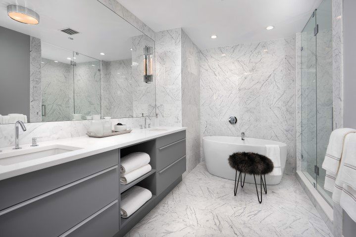 Modern bathroom with grey vanity, white marble surfaces, freestanding tub, glass shower, and a stool with a dark cushion. Towels are neatly placed on the shelves and rack.