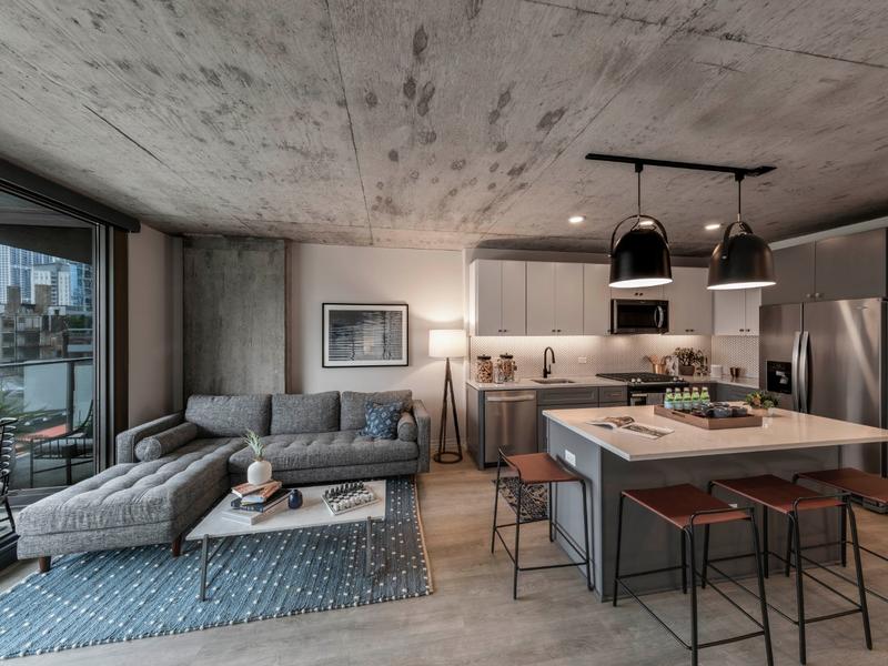 Modern open-plan apartment with a gray sectional sofa, a white coffee table, a kitchen island with stools, pendant lights, and a large window. Concrete ceiling and wooden floors are visible.