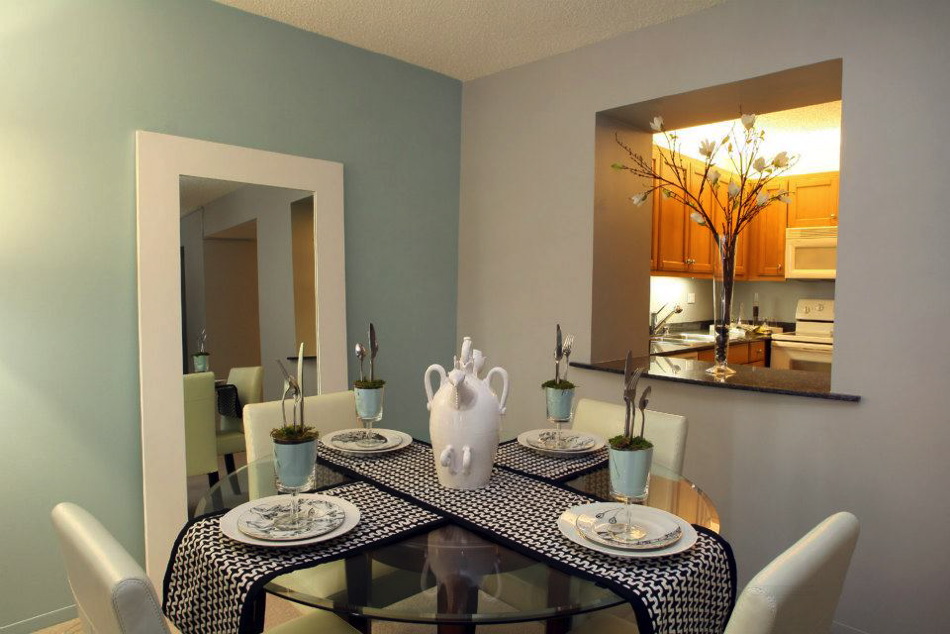 Dining area with a glass table set for four, featuring plates and a decorative vase. A large mirror reflects the space. Kitchen with wooden cabinets is visible in the background.