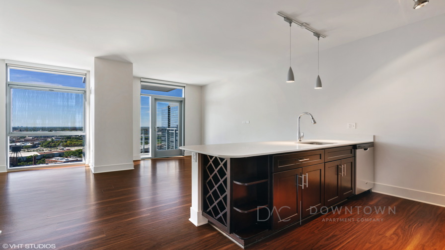 Modern open-concept apartment with hardwood floors, large windows, an island with built-in wine rack, and pendant lighting above.