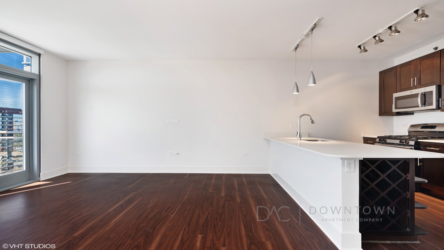 Modern apartment with an open floor plan, featuring a kitchen with dark cabinetry, stainless steel appliances, pendant lights, and a bright living area with wood flooring and large windows.