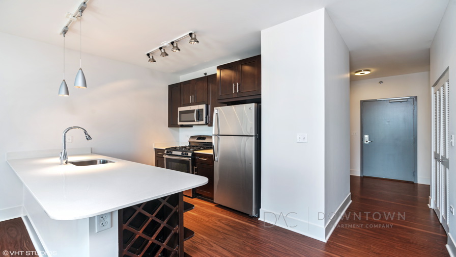 Modern kitchen with stainless steel appliances, dark wood cabinets, and a white countertop under pendant lights. Wood flooring extends to an adjacent hallway with a closed door.