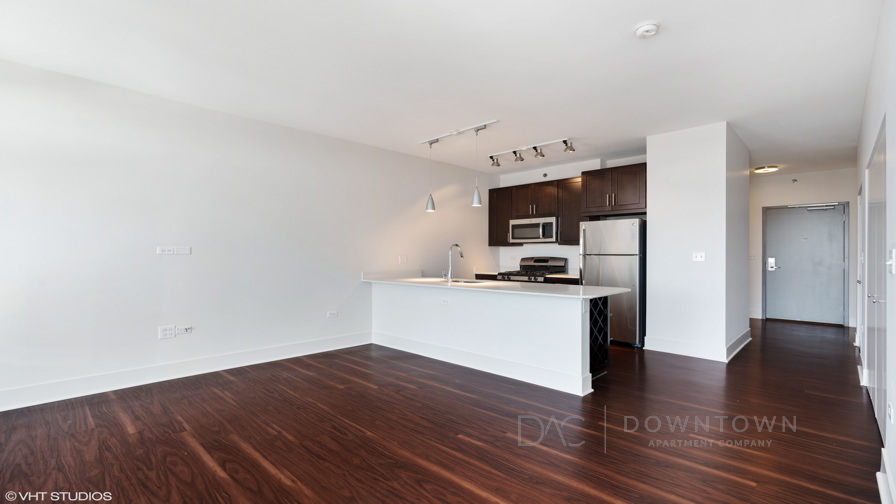 Modern kitchen with stainless steel appliances, dark wood cabinets, and an island with pendant lights. Hardwood flooring and white walls complete the open space.