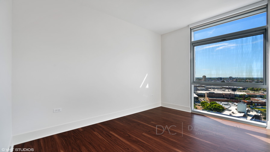 An empty room with wooden flooring, a large window, and a view of a cityscape under a clear blue sky.