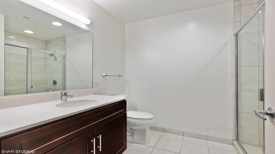 Modern bathroom with a large mirror, white countertop, dark wooden cabinets, toilet, and a glass-enclosed shower.